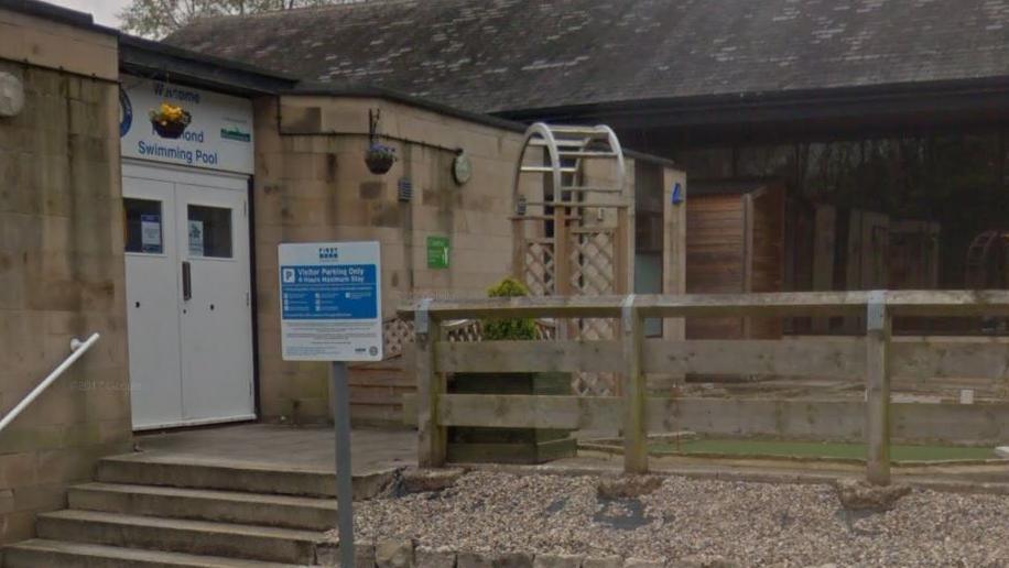 Two white doors and a hanging flower basket . Above the doors in blue lettering it says Richmond Swimming Pool. In the front is a wooden fence.