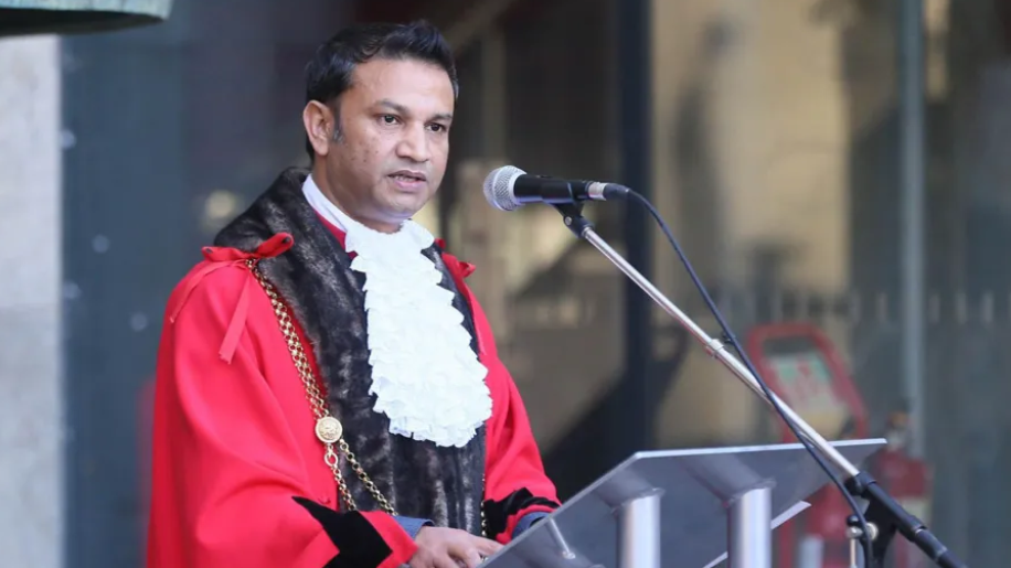 A man in a red robe with brown fur trim and white ruffled shirt standing in front of a microphone, giving a speech.