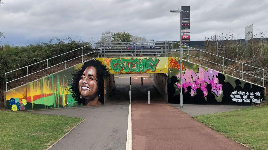 A colourful mural on the end of an underpass, showing a smiling woman on the right with her face raised, Gateway written above its entry in green letters and figures silhouetted in black against pink letters on the right, beneath the A10 with a car midway across the bridge