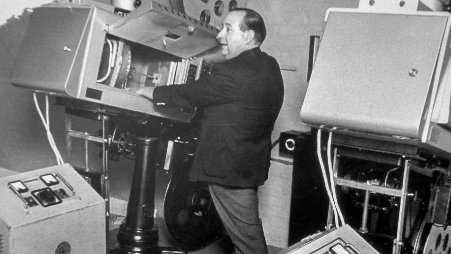 A black and white photo of Colin Aldis wearing a suit and putting some film reel into a large silver machine. He has short hair and is surrounded by various film equipment.