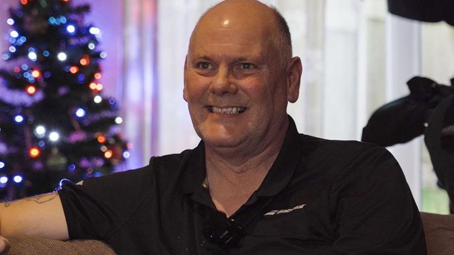 Nigel in a black t-shirt sitting on his sofa and smiling broadly. There is a Christmas tree in the background with lights. 