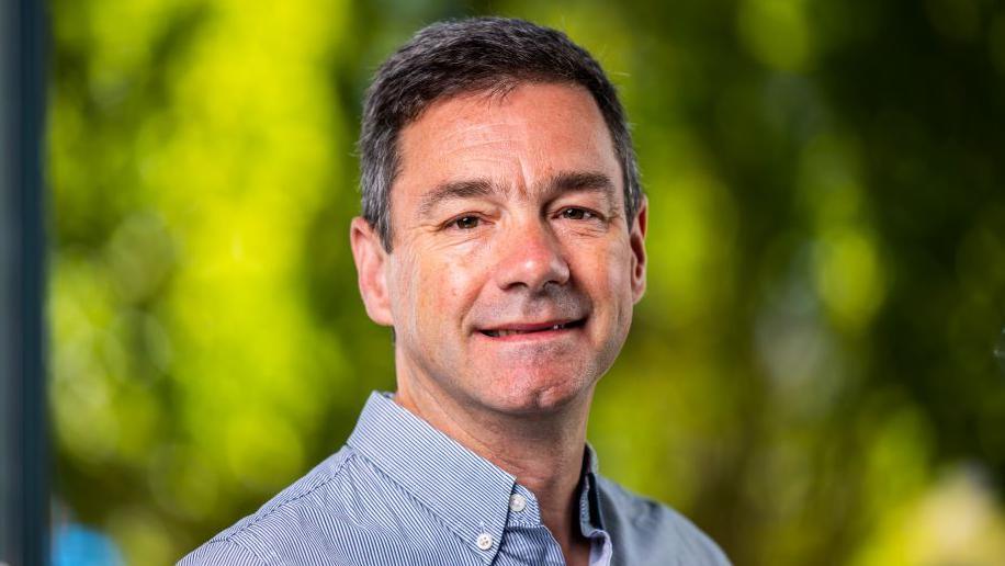 Lawrence Gosden smiling at the camera is wearing a blue collared shirt, has dark hair and eyes. Green foliage in the background is blurred.