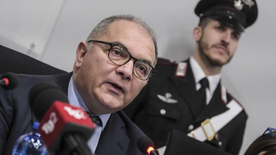 An anti-mafia prosecutor wearing glasses sits in front of a red microphone, as a member of the Italian Carabinieri stands behind him