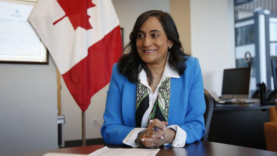 Anita Anand, during an interview in her office in Ottawa, wearing a blue jacket and a patterned scarf. She is sitting at a desk and a Canada flag hangs behind her