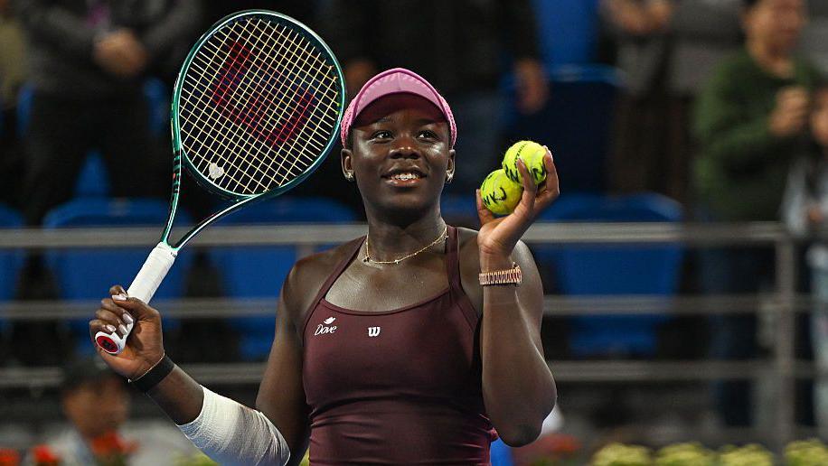 Victoria Mboko plays a backhand. She has black hair, tied back, and is wearing a pink cap and maroon dress. Her right arm is bandaged.