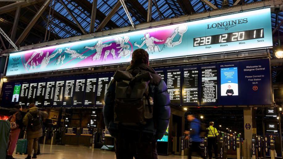 A man with a backpack standing in a railway station, looking up a the large departure boards and the countdown clock of the Commonwealth Games - a blue and purple designed illustration of athletes playing sport, with a clock ticking down to the Games starting. 