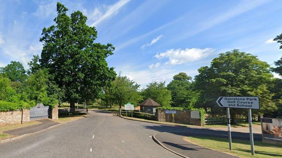 Entrance to Overstone Park, with resort sign to the left, road sign to the right and trees flanking the entrance