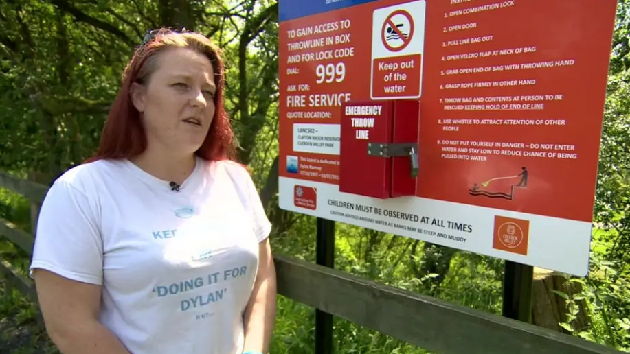 Beckie Ramsay, wearing a white 'Doing It For Dylan' t-shirt, stands in front of a safety warning sign near open water