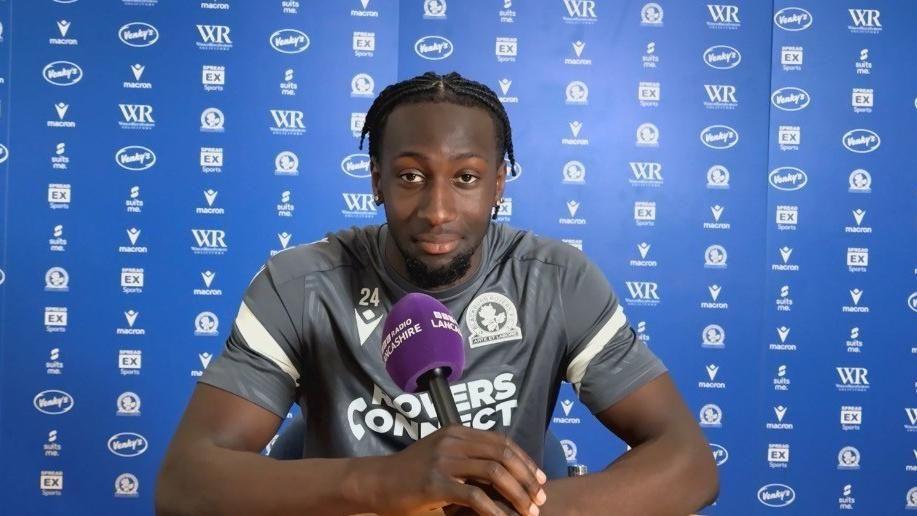 Moussa Baradji is sitting with a BBC microphone infront of him with his arms leaning on a desk with a sponsor background behind him. He is wearing a Blackburn Rovers grey training top with the number 24 on it.