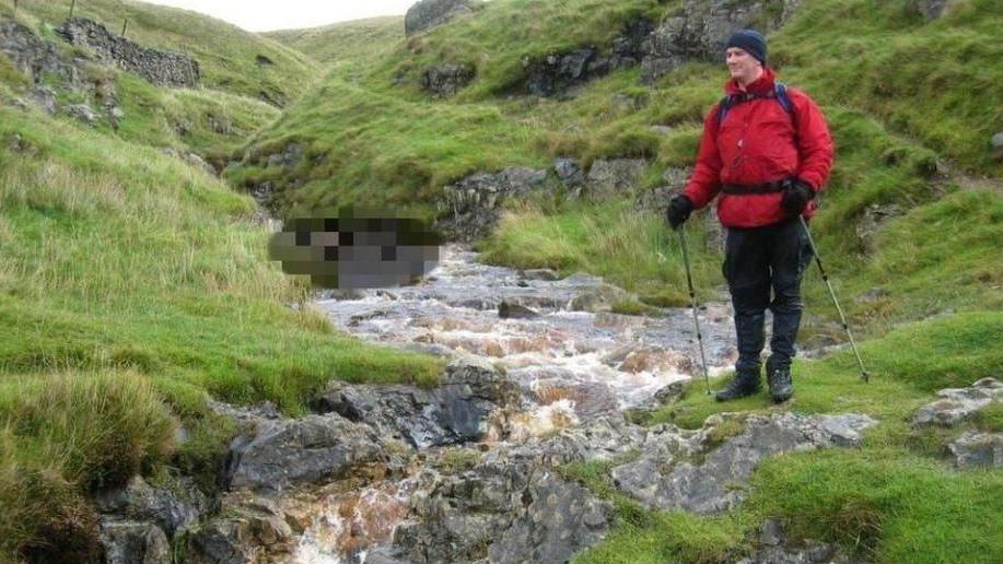 A man posing next to a rocky stream running through hills in the Yorkshire Dales. The man is wearing a red jacket, black trousers, a hat and gloves and is holding walking sticks.