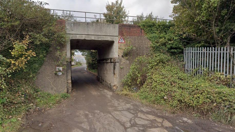 Walnut Tree Bridge, a narrow concrete rail bridge over a single track road, with slightly overgrown foliage on either side and a grey metal fence to the right.