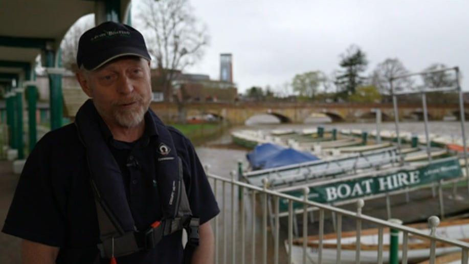 A man speaking by a river with boats in the background