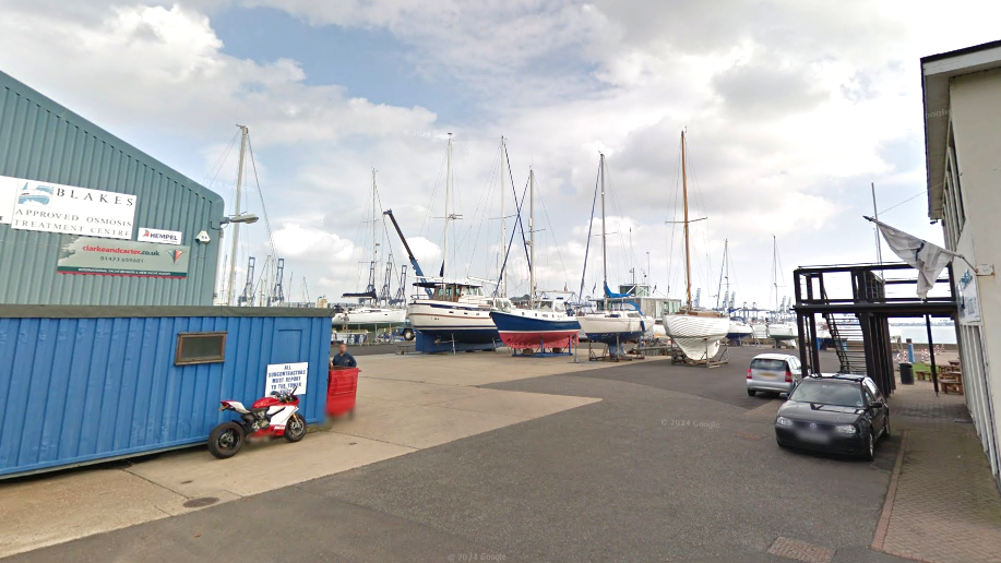 The forecourt and parking area of a marina. There is an industrial building painted blue and turquoise to the left. In the background are sail boats. There are a couple parked cars. There is some blue sky and white clouds above.