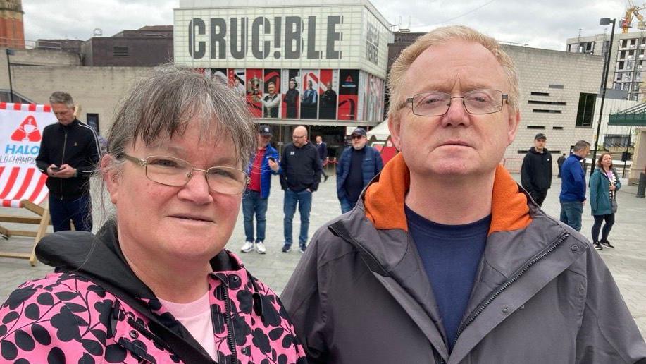 A woman with hair tied back and glasses wearing a patterned pink and black coat stands next to a middle-aged man with glasses wearing a grey coat.