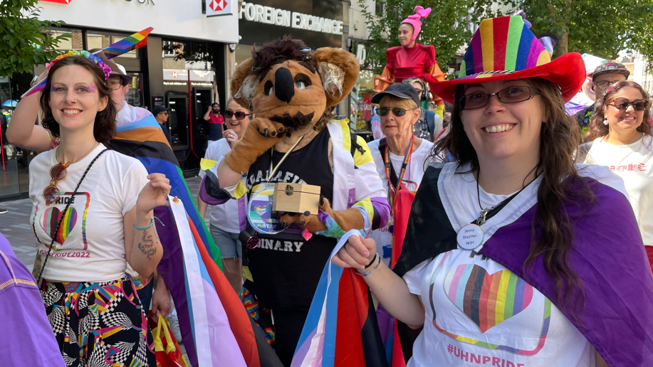 A group of people on the pride walk are heading towards the camera. There are two women in front wearing white tops with #UHN pride written on the front with a love heart - coloured in white, pink, light blue, brown, black, red, orange, yellow, green, dark blue, and a darker pink. They are smiling.
