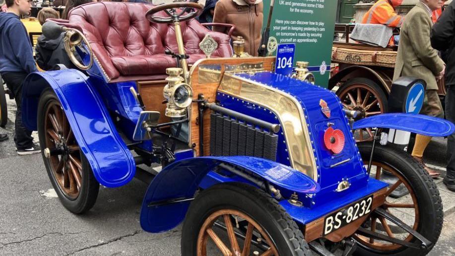 A royal blue 1902 Deckert two-seater open-top car. Its wheels and steering wheel are wooden, while the seat is made of a plush-looking red leather.