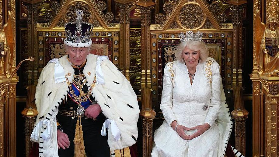King Charles III sits besides Queen Camilla during the State Opening of Parliament in the House of Lords, both are on thrones and wearing full regalia and crowns 