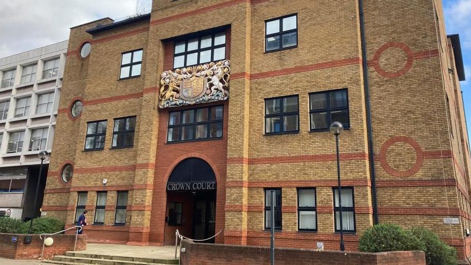 The outside of St Albans Court Court, showing a brick building, with a courts of justice coat of arms. with black windows, a large entrance and steps up to the building. 