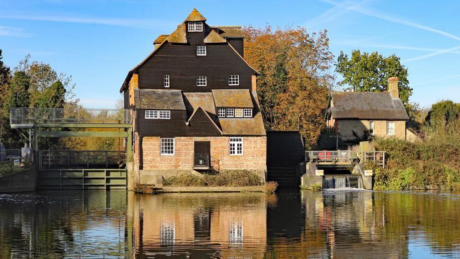 An external shot of Houghton Mill, a stone building on a river with the watermill to the right.