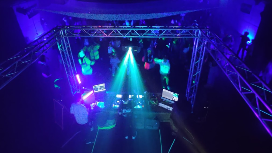 An aerial shot of a darkened nightclub, with the back of the DJ and their equipment in the foreground, and behind, people dancing under purple and green lights