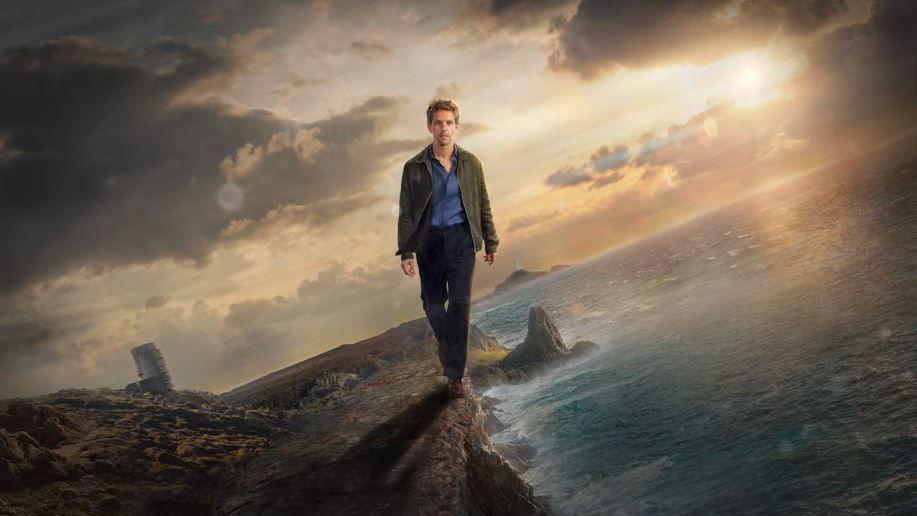 Damien Molony as Det Jim Bergerac walking along a cliff with the sea to his left and Jersey to his right. The sky is cloudy and the sun is setting behind him. He is wearing a green jacket with a blue shirt and black trousers.