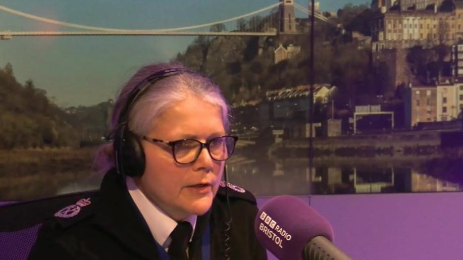 The Chief Constable of Avon and Somerset Police Sarah Crew is pictured in her uniform sat in the BBC Radio Bristol studio, wearing black headphones talking into a purple BBC microphone.