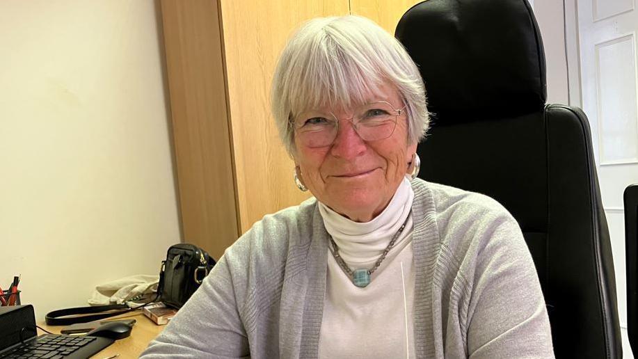 A woman with grey hair in a white top and pale grey cardigan, sitting on a high-backed black chair at a wooden desk with a black keyboard on it