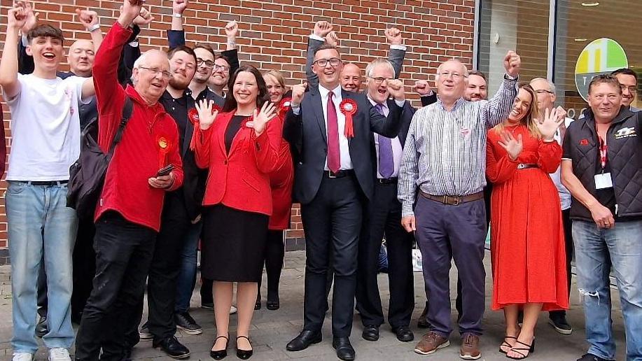 Labour MP Connor Naismith, wearing a dark blue suit and red rosette, celebrates with numerous supporters dressed in red 