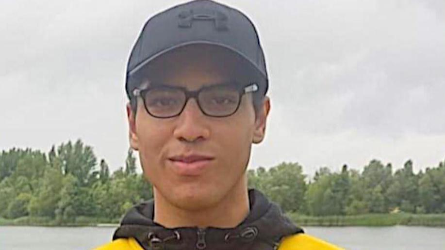 Nicholas Prosper dressed in glasses, a black baseball hat and yellow and black hoodie while standing in front of a lake.