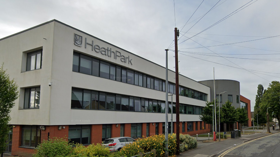 The school building is grey and red brick and sit opposite houses with some shrubbery in front of it.