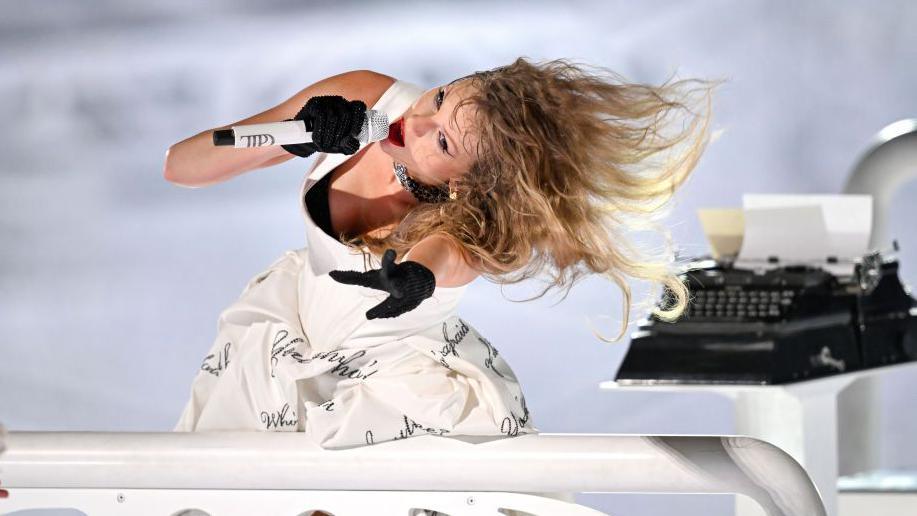 Taylor Swift wearing a white dress sings on stage with a typewriter behind her.
