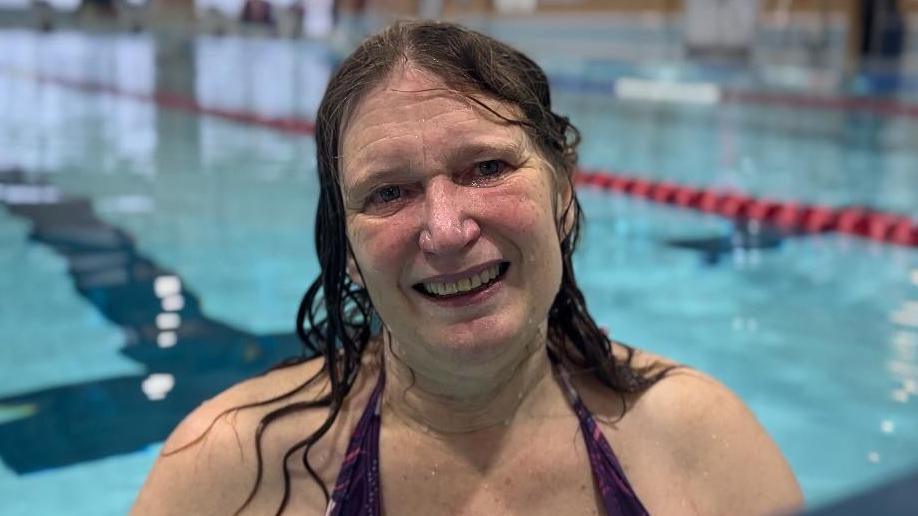 A woman with long, dark, wet hair. She is looking and smiling at the camera while standing in the water by the edge of a pool.