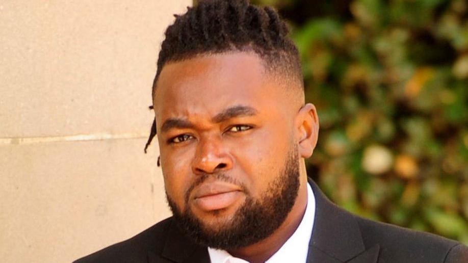 A man with a beard and dark hair, closely cropped at the sides, wearing a suit, pictured outside a court