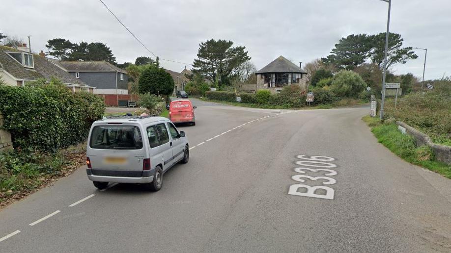 The Consols junction in St Ives. Two vehicles - a silver Peugeot car and a red Ford van - are turning left at the junction. It is a cloudy day and there are houses on the left hand side along with a building on the corner of the junction.