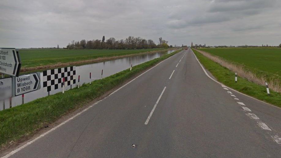 A long, straight rural road runs alongside a water-filled ditch. There are road markings for a junction to the right and road signs to the left.