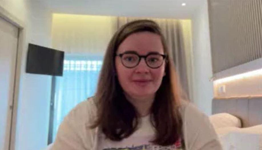 Aine McMenamin, who is wearing glasses and a T-shirt and has mid-length brown hair, sits in a small white hotel room with a TV on the wall.