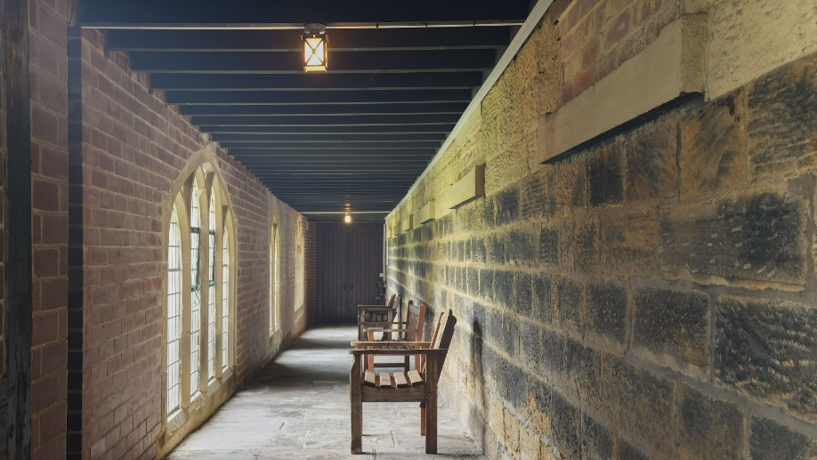 A corridor in a monastery with three benches. There is original brick stonework on the right-hand side and newer brickwork to the left. There are three lots of arched windows and black beams across the ceiling.