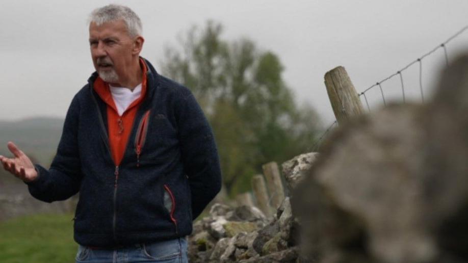 A picture of a man standing in a field wearing a blue jacket with a red top underneath and blue jeans. 