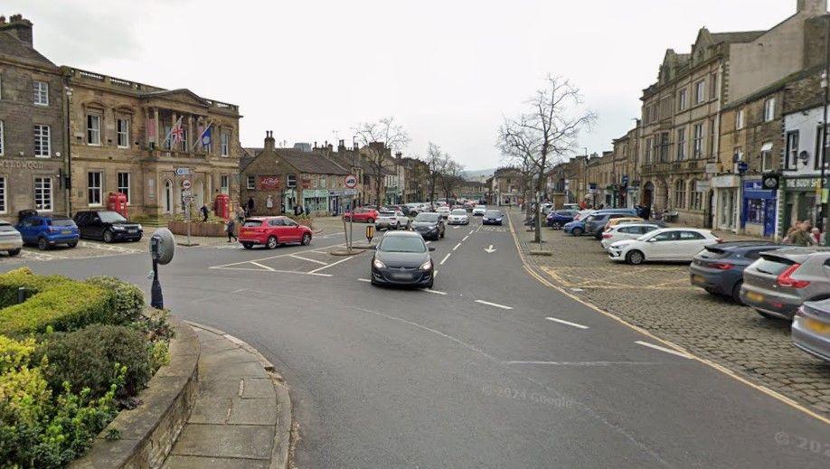 A busy roundabout in a market town. Parked and moving cars can be seen.