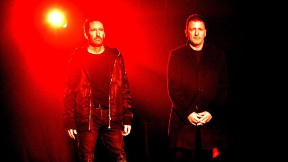 Nine Inch Nails members Trent Reznor and Atticus Ross stand against a dark background, illuminated by a red light.