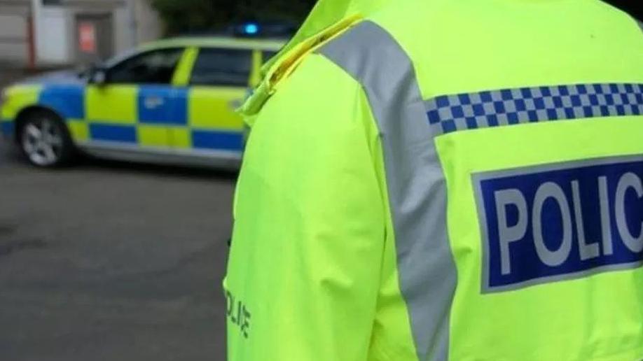 A police officer standing to the right in a hi-vis jacket with police written on the back