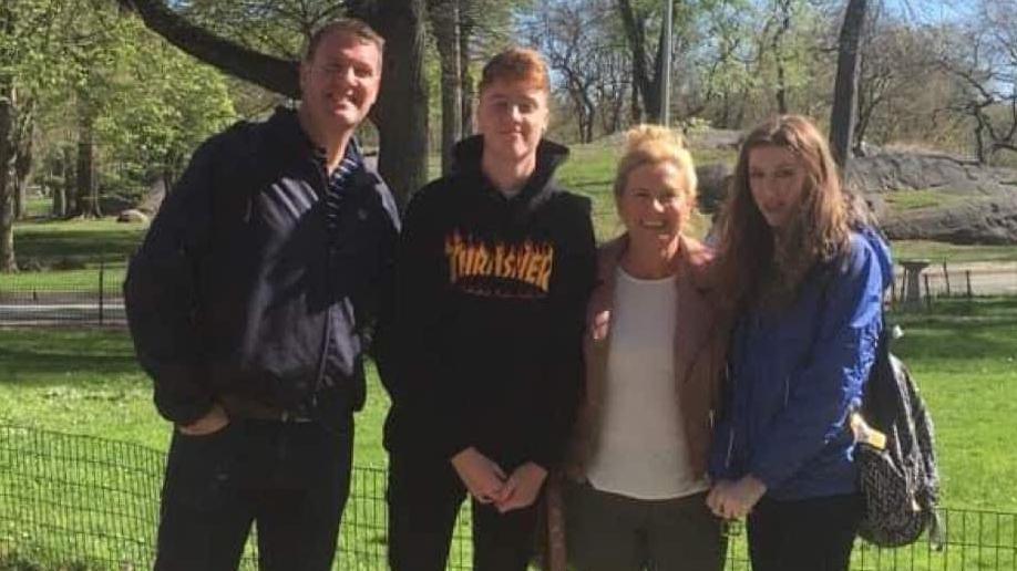 Steven Linton, Tom Linton and Amanda Linton are smiling at the camera while they have their photo taken at a park. Steven Linton has short brown hair and is wearing black trousers and black jacket. Tom Linton is wearing a black hoodie and black trousers. Amanda Linton's blonde hair is tied back in a bun. She is wearing a white top, pale pink jacket and grey trousers.