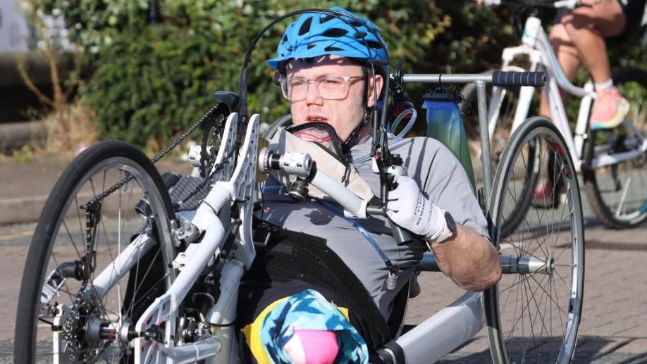 A man wearing glasses, a blue helmet and a grey top is pictured riding a three-wheeled recumbent bike.