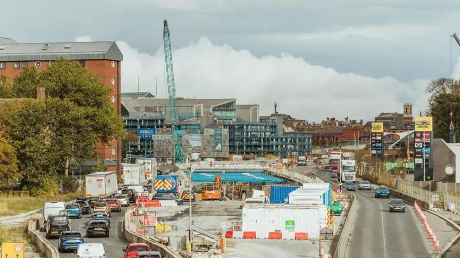 A view of a large roadworks project in the centre of a busy dual-carriageway, which splits around the work area. Cranes, diggers and temporary buildings can be seen. The buildings of Hull city centre can be seen in the background.