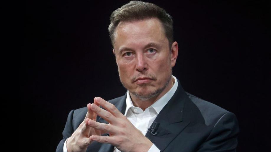 Elon Musk, who owned social media platform X, pictured with a stern expression, with his hands put together. He is dressed in a dark grey suit against a black backdrop.