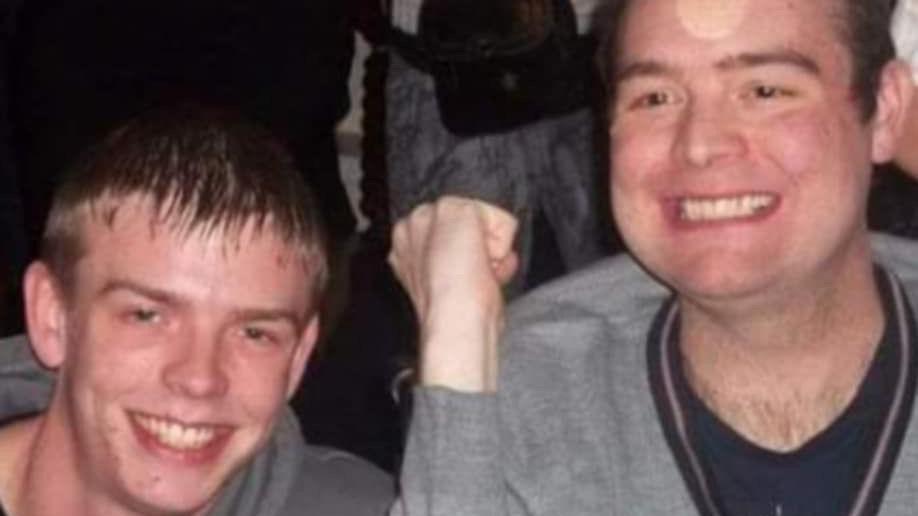 Flash photograph of two young men, both wearing grey hoodies and dark T-shirts. Toby, on the left of the photo, is smiling and looking straight into the camera. Ben, on the right, is smiling and looking slightly off to his right, with one fist slightly raised.