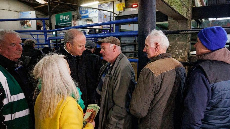 Micheál Martin, who has balding light brown hair and is wearing a dark coat talks with three farmers at a livestock market. The man closest to him is wearing a flat cap and green coat, while the man behind him has grey hair and is wearing a brown coat.