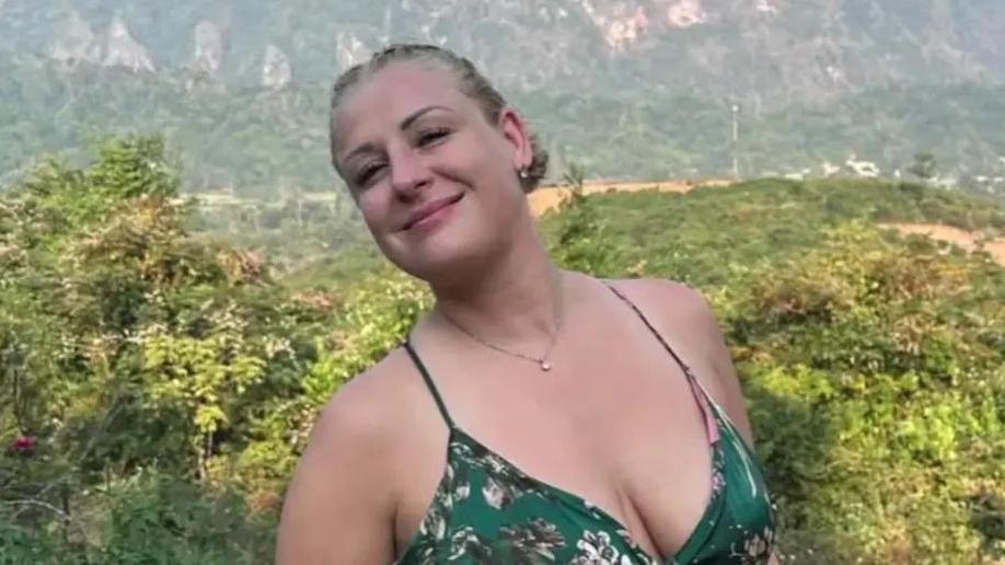A blonde woman wearing a green dress. She is smiling and there are green trees and hills in the background.