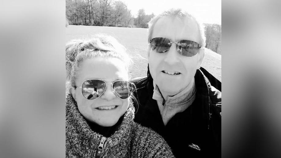 A man and woman pictured outdoors, smiling at the camera. Both are wearing sunglasses. The woman has her hair pulled back and is dressed in a jumper. The man is wearing a jacket with the brand name 'Trespass' visible. Behind them is an open field with trees in the distance.