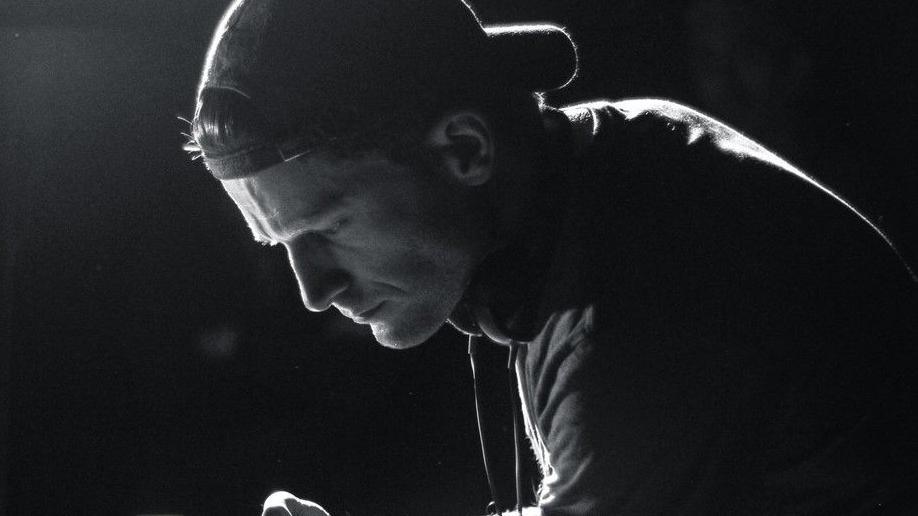 A side profile of Arthur's face in black and white, silhouetted by light. He is leaning over a camera wearing a baseball cap backwards
